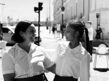 Two women in white shirts, facing each other and laughing, on a sunlit street